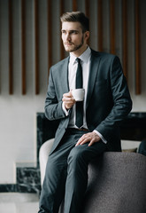 Wall Mural - Young handsome man sitting in office with cup of coffee and working on project connected with modern cyber technologies. Businessman with notebook trying to keep deadline in digital marketing sphere.