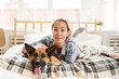 Portrait of smiling Asian woman hugging dog lying in bed and looking at camera, copy space