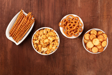 Wall Mural - Salty crackers, sticks, pretzels, and goldfishes, shot from the top on a dark wooden background with copy space. Party snacks mix with a place for text