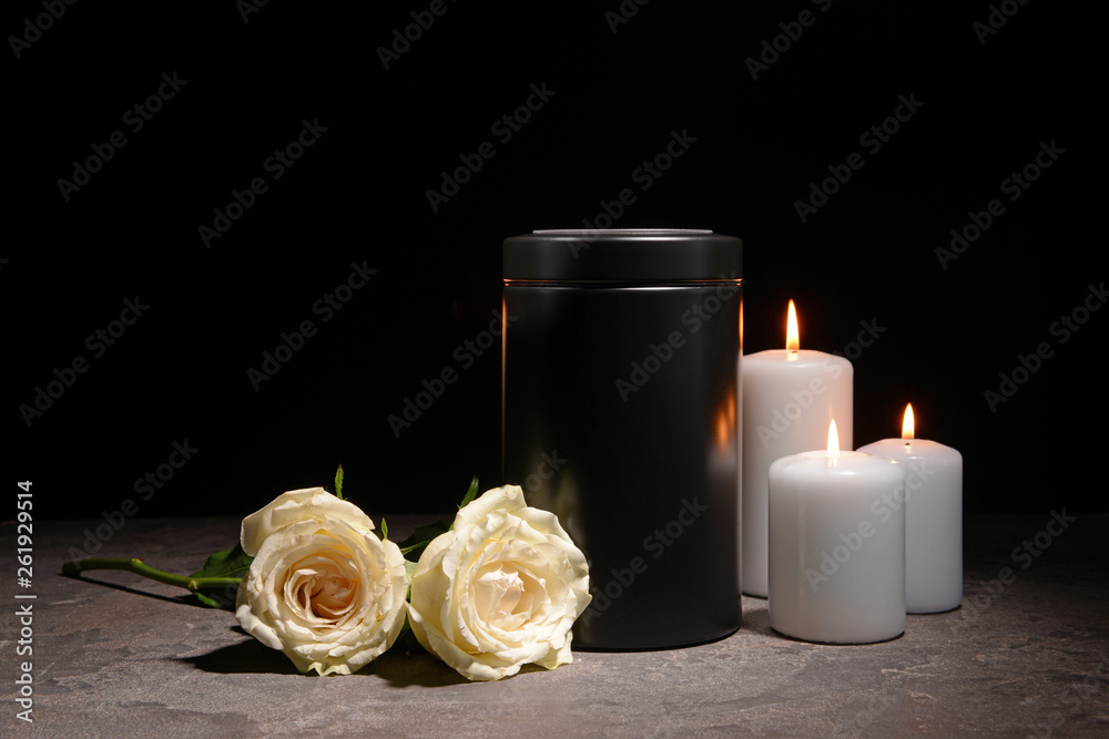 Mortuary urn, burning candles and flowers on table against dark background