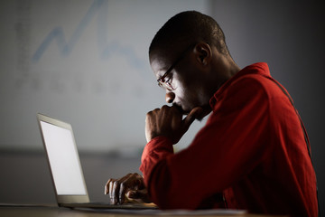 Side view portrait of thoughtful African businessman looking at laptop screen while working in dark office, copy space