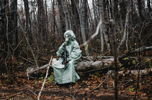 Stalker, A Man In A Gas Mask And Special Chemical Protection, Green Cloak.  With A Gun In His Hand, He Sits On A Stump In The Forest.