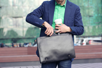 Cropped image businessman in casual wear with cup of coffee to go puts smartphone into trendy leather briefcase against modern office building background