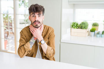 Poster - Young man wearing casual jacket sitting on white table shouting and suffocate because painful strangle. Health problem. Asphyxiate and suicide concept.