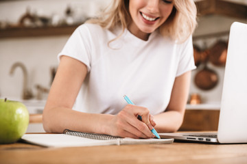 Poster - Image of cute blond woman 20s wearing white t-shirt studying on laptop and writing down notes in apartment