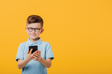 handsome little boy is happy with the phone in hand, in glasses, dressed in blue shirt, isolated on yellow background, copy space