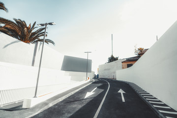 A two-lane modern asphalt road, resting against the gate with the barrier, two whites walls, arrows as the road marking, palm and streetlights on the left, superbright warm day, Lisbon, Portugal