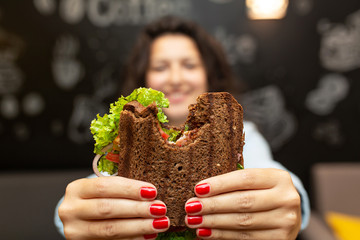 Wall Mural - Closeup funny blurred protrait of young woman hold bitten sandwich by her two hands. Sandwich in focus. dark background