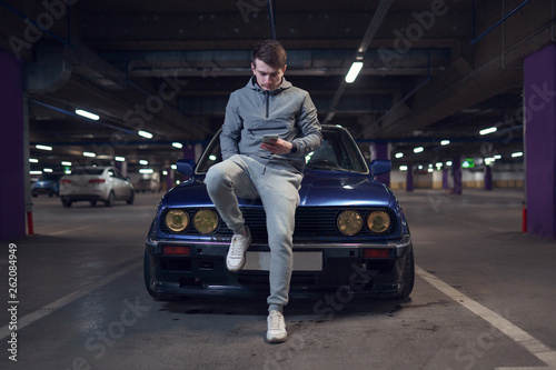 Fotomural Photo of man with phone sitting on hood of black car.