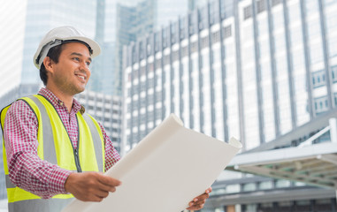 Young asian engineer holding layout plan of building and smile on construction site background. Construction manager concept.