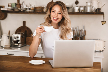 Wall Mural - Image of cute blond woman drinking tea and using laptop while sitting at wooden table in apartment