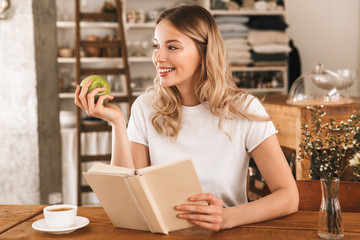 Wall Mural - Portrait of smart blond woman reading book and eating green apple while sitting in cozy cafe indoor