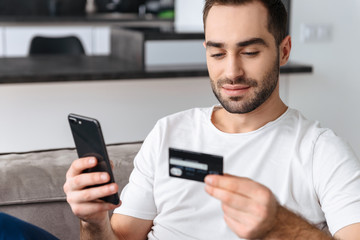 Sticker - Photo of caucasian guy using smartphone and holding credit card while sitting on sofa at home