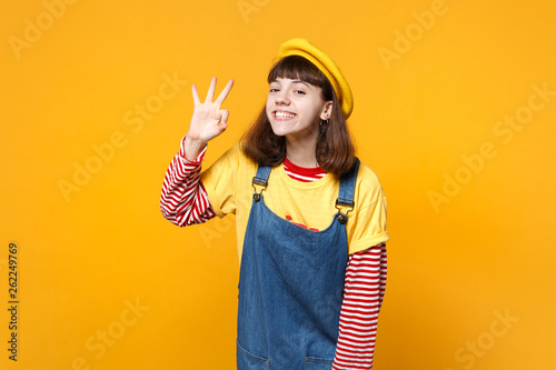 Cuadros en Lienzo Portrait of smiling girl teenager in french beret, denim sundress showing OK gesture isolated on yellow wall background in studio