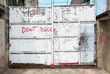 White colored old rusty metal entrance gate, closed and locked and secured with barbed wire. 'Don't Block' is written on the gate.
