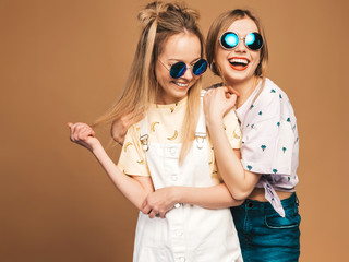 Two young beautiful smiling blond hipster girls in trendy summer colorful T-shirt clothes. Sexy carefree women posing near beige wall in round sunglasses. Positive models having fun