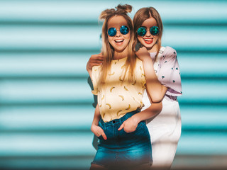 Two young beautiful smiling blond hipster girls in trendy summer colorful T-shirt clothes. Sexy carefree women posing near blue wall in round sunglasses. Positive models having fun