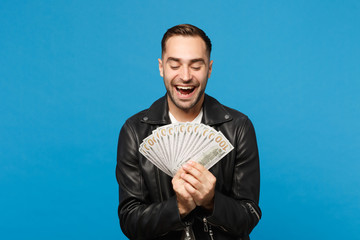 Young unshaven man in black leather jacket white t-shirt holding fan of cash money in dollar banknotes isolated on blue wall background studio portrait. People lifestyle concept. Mock up copy space.