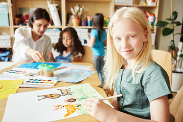 Adorable albino girl smiling and looking at camera while sitting at table and painting nice picture during lesson in art school