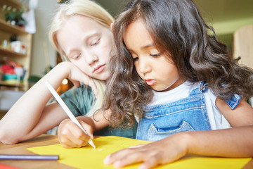 Two charming little girls sitting at desk and drawing picture on yellow paper while studying in art school together