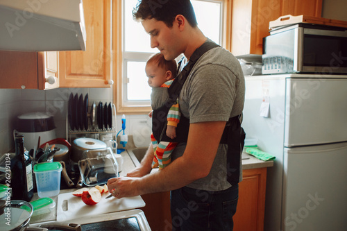 Young Caucasian father with newborn baby in carrier preparing lunch. Man parent carrying child standing in kitchen making food. Authentic lifestyle candid real moment. Single dad family life concept
