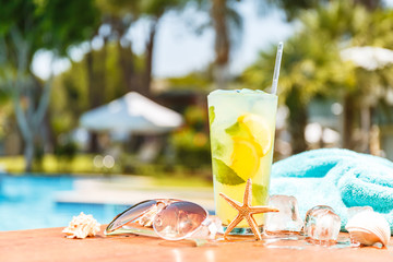 Wall Mural - Glass of classic mojito cocktail and caipirinha with seashells, sunglasses and blue towel closed to swimming pool. Vacation concept.