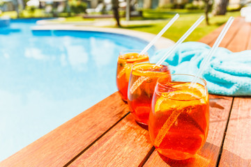 Wall Mural - Three glasses of aperol spritz and negroni cold cocktail on wooden edge of swimming pool with turquoise towel. Vacation concept. Place for text.