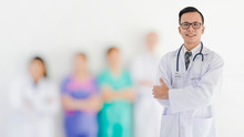 Portrait Of Smiling Asian Medical Male Doctor Standing In Front Of His Blurred Team Staff In Hospital Background.Concept Of Effective Work In Hospitals.