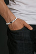 Cropped half-turn shot of guy's hand with stony marbled bracelet with black and white stones. The man in black jeans and white T-shirt is putting his hand into a pocket, posing on dark background.