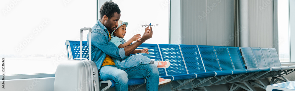 panoramic shot of african american father and son sitting with suitcase in airport and playing with toy plane - obrazy, fototapety, plakaty 