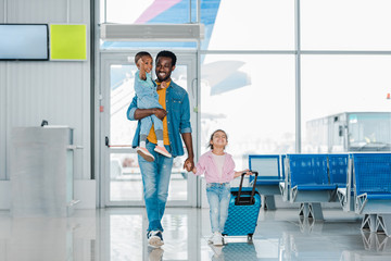 smiling african american father walking with happy children and baggage along waiting hall in airport