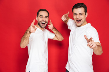 Image of two beautiful men friends 30s in white t-shirts smiling and pointing fingers at camera