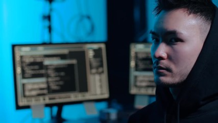Poster - cybercrime, hacking and technology concept - young asian hacker taking his hood off in dark room with computers at night
