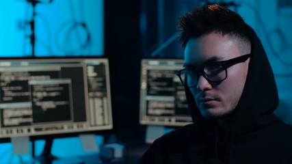 Poster - cybercrime, hacking and technology concept - young asian hacker wearing glasses and hoodie in dark room with computers at night