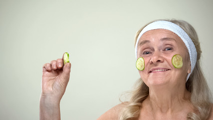 Wall Mural - Funny joyful granny with cucumber mask on face smiling on camera, cosmetology