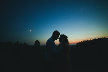 Silhouettes Of A Young Couple On A Sunset Background, The Moon In The Sky. The Bride And Groom Stand Against The Setting Sun. The Guy And The Girl Against The Sky At Sunset. The Concept Of Love.