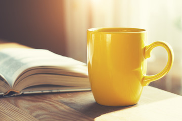 Wall Mural - yellow coffee cup and open book on wooden table, morning sunlight background