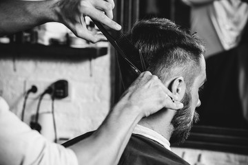 Young handsome barber making haircut for attractive bearded man at barbershop. Black and white or colorless photo. Hairstyle, salon, hairdresser, barber shop, lifestyle concept. Caucasian male models.