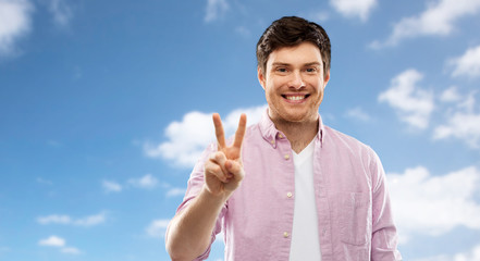 gesture, count and people concept - smiling young man showing two fingers or peace hand sign over blue sky and clouds background