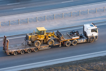 Truck with a long trailer platform for transporting heavy machinery, loaded tractor with a bucket. Highway transportation.