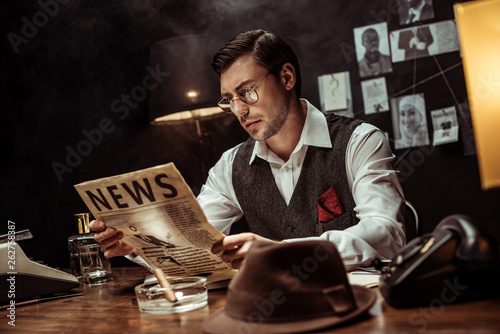 Concentrated detective in glasses reading newspaper in dark office Poster Mural XXL