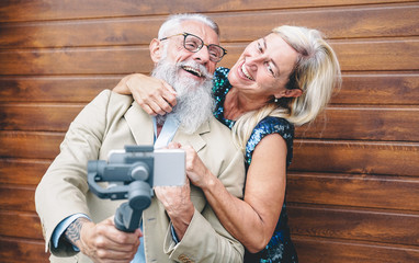 Happy seniors couple recording video with gimbal mobile phone outdoor - Mature fashion people having fun with new trends smartphone apps - wooden background - Elderly lifestyle and technology concept