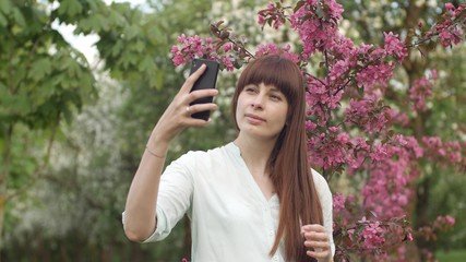 Girl makes selfie in the garden. An attractive red-haired woman smiles making selfi using a mobile phone in a cherry orchard. The concept of using gadgets for a healthy lifestyle.