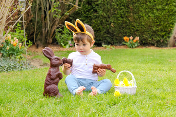 Wall Mural - Cute little baby boy with bunny ears on head holding  chocolate Easter bunnies sitting on green grass outside in the spring garden with basket of easter eggs looking at big chocolate rabbit. 