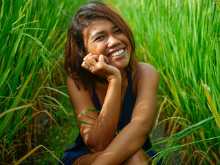 natural and fresh portrait of young happy and exotic islander Asian girl from Indonesia smiling cheerful and excited posing in green field playing with rice enjoying holidays