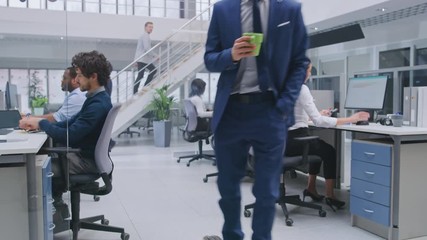 Poster - Young Happy Handsome Manager Wearing a Suit and Tie Rides a Longboard. He's Holding a Cup of Coffee. Diverse and Motivated Business People Work on Computers in Modern Open Office.