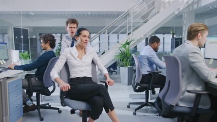 Poster - Cheerful Employee Pushes His Beautiful Female Colleague on a Chair Between Rows of Desks with Diverse Business People Working on Desktop Computers in Modern Office Space.