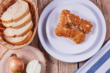 Deep-fried battered cod fillets, in a white plate next to onions and bread on a wooden background. Flat lay