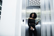 © AS Photo Family - Fashionable young beautiful african american business woman with afro hairstyle wear in elegant black, stand at elevator with phone and laptop at hands.