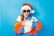 © Alex - A young girl in a Santa Claus hat is holding gift boxes on a blue background. The concept of New Year and Christmas, gifts for the winter holidays, shopping at sales.